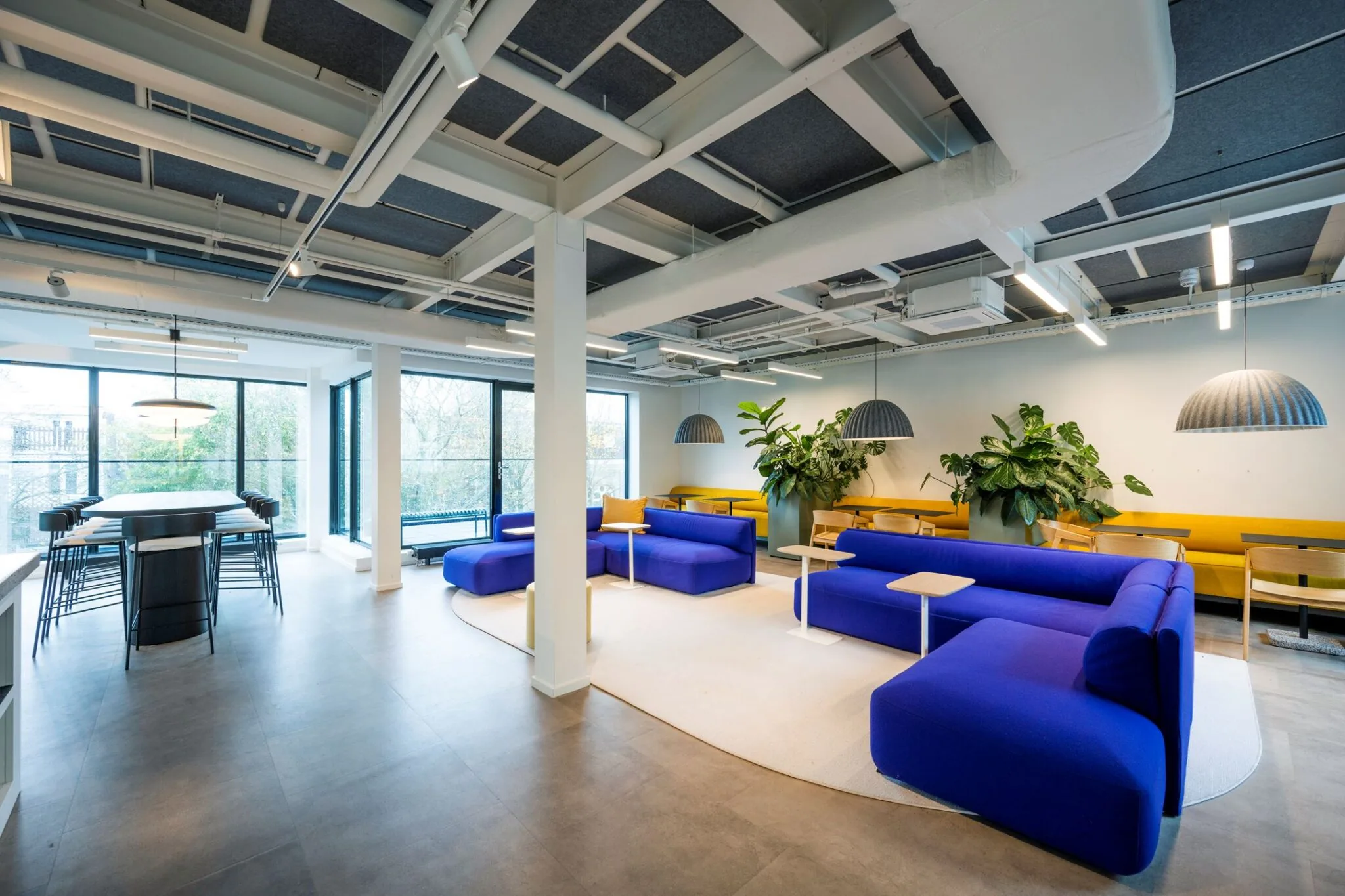 Moderne lounge- en werkruimte op Keizersgracht 572 met blauwe banken, gele zitbanken, grote ramen en veel groen.