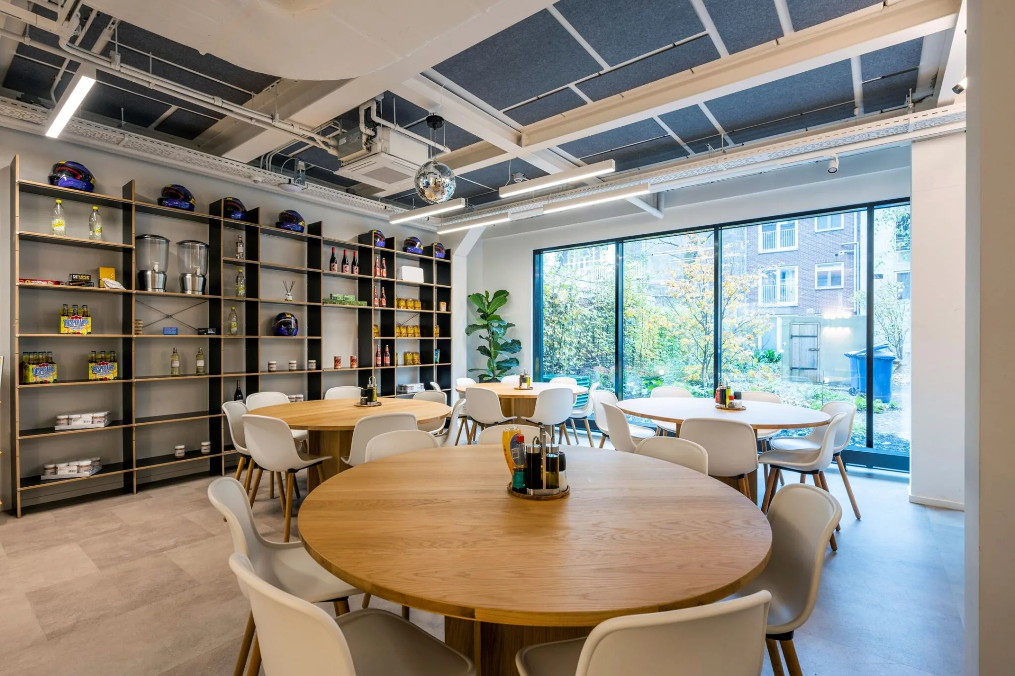 Moderne kantineruimte aan de Keizersgracht 572 met ronde houten tafels, witte stoelen, een grote wandkast en uitzicht op een groene binnentuin.