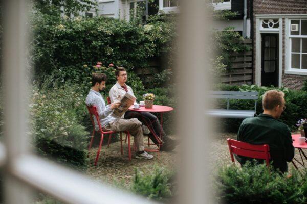 Drie mannen zitten aan rode tafels in een groene tuin bij Keizersgracht 64, terwijl één van hen een krant leest.