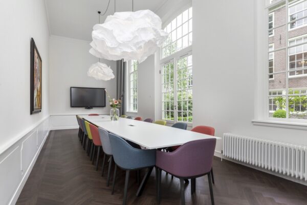 Vergaderruimte in Keizersgracht 64 met lange witte tafel, kleurrijke stoelen, grote ramen en speelse hanglampen.