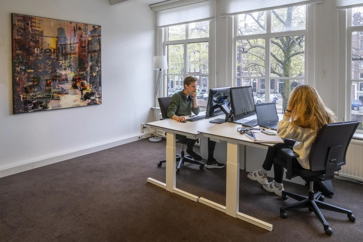 Twee mensen werken aan bureaus met computers in een licht kantoor aan de Keizersgracht, met kunst aan de muur en uitzicht op de gracht.