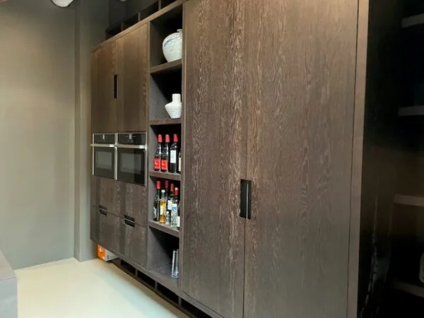 Donkere houten keukenkasten met ingebouwde ovens en open schappen met flessen en servies.