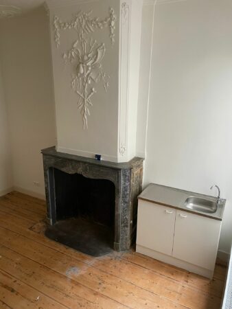 Een klassieke schouw met sierlijk stucwerk naast een eenvoudig wit keukenkastje met spoelbak in een kamer aan de Kloveniersburgwal 56.