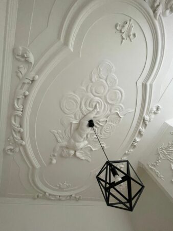 Plafondornament met sierlijke stucwerkdecoratie en engelfiguur aan het plafond van Kloveniersburgwal 56, met een moderne zwarte hanglamp.