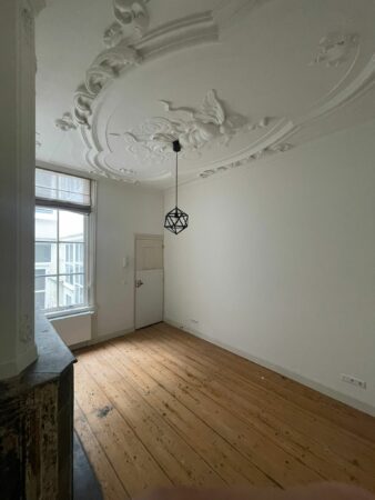 Lege kamer in Kloveniersburgwal 56 met houten vloer, sierlijk stucplafond en moderne hanglamp.