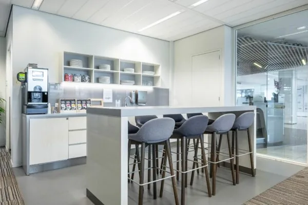 Moderne kantoorkeuken met een hoge tafel, barkrukken, koffiemachine en open kastruimte op Koningin Juliana Plein 10.