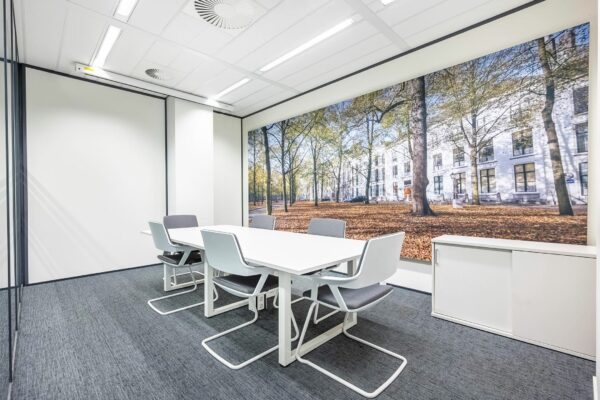 Moderne vergaderruimte met witte tafel, grijze stoelen en een grote wandfoto van het Koningin Julianaplein in de herfst.