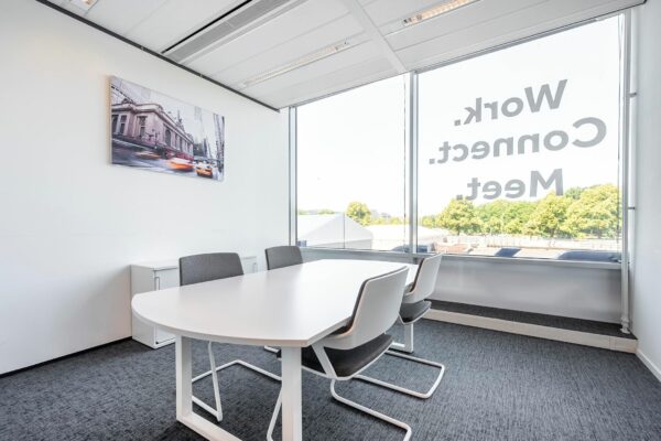 Moderne vergaderruimte aan het Koningin Julianaplein met witte tafel, grijze stoelen en groot raam met de tekst "Work. Connect. Meet."