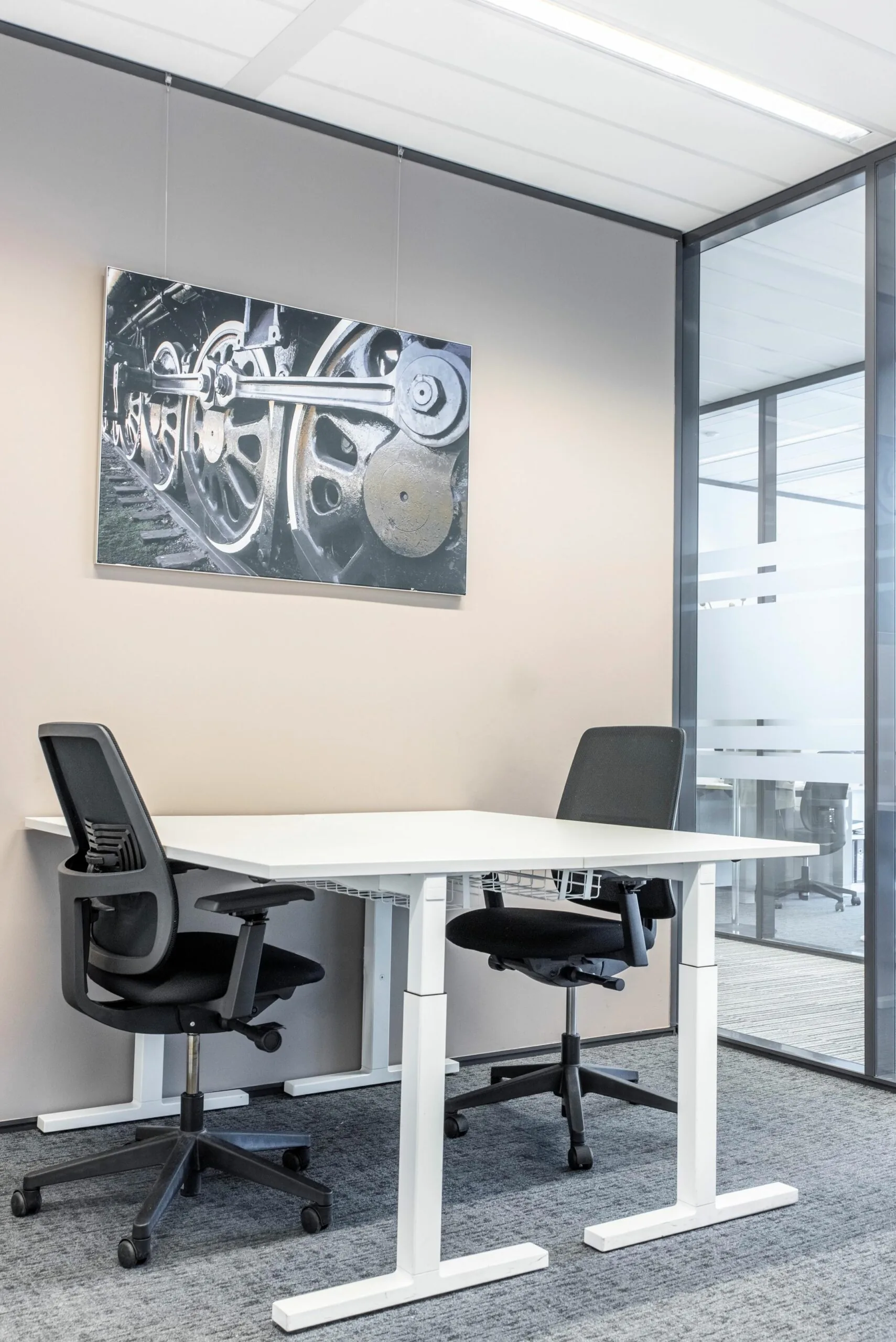 Kleine vergaderruimte op Koningin Juliana Plein 10 met twee bureaustoelen, een witte tafel en een foto van treinwielen aan de muur.