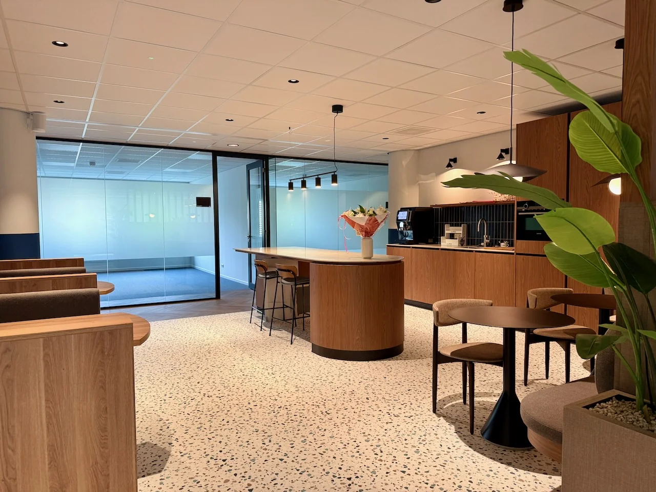 Moderne loungehoek in Kronenburg Offices met houten zitjes, ronde tafels, een koffiehoek en grote kamerplant.