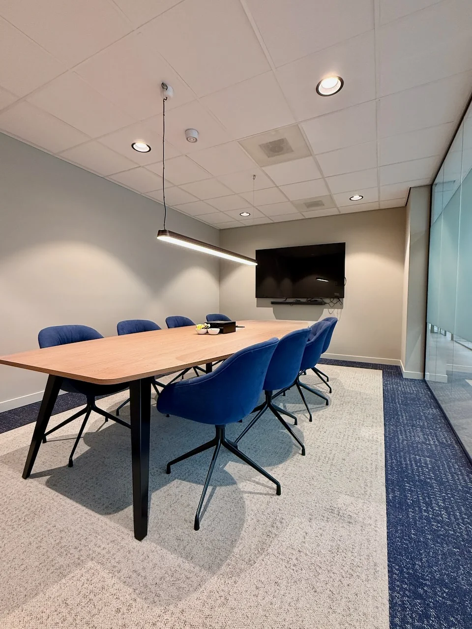 Vergaderruimte in Kronenburg Offices met een houten tafel, zes blauwe stoelen, een hanglamp en een flatscreen aan de muur.