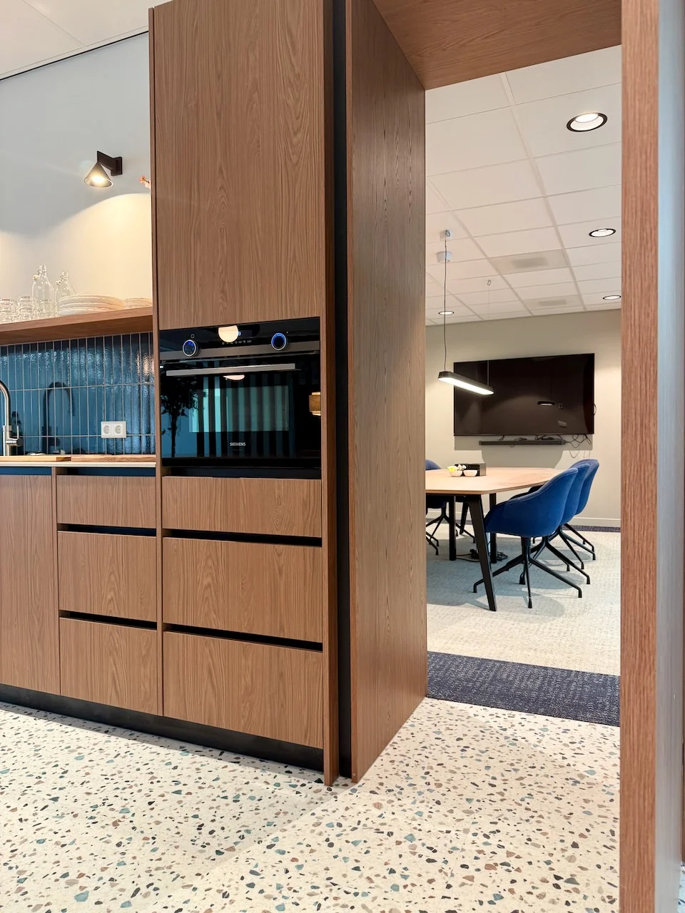 Moderne kantoorruimte Kronenburg Offices Meet 2 met houten kitchenette en vergaderruimte met blauwe stoelen op de achtergrond.