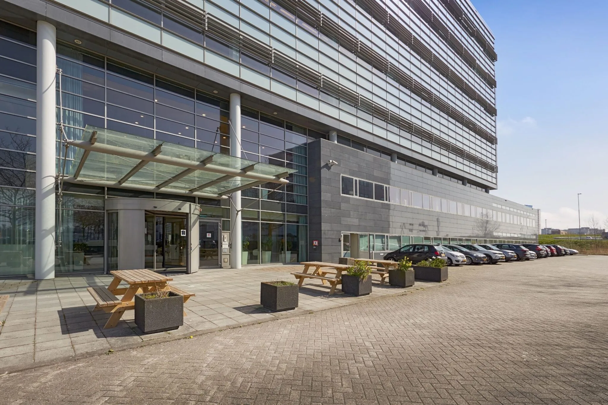 Ingang van het kantoorgebouw aan de Laarderhoogtweg 7 met glazen gevel, parkeerplaatsen en picknicktafels.