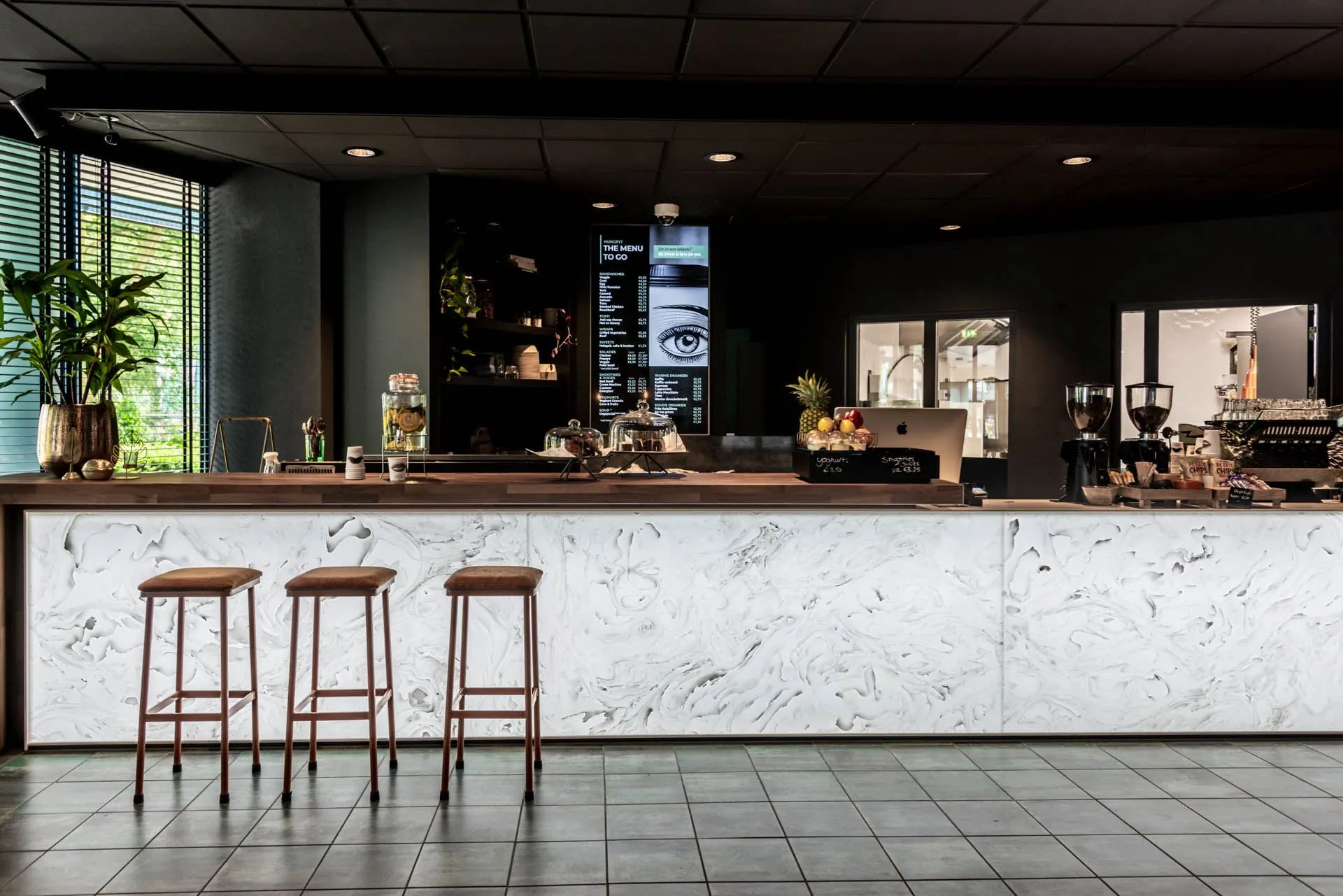 Moderne koffie- en lunchbar met marmeren toonbank, drie barkrukken en een digitaal menu aan de muur bij Lange Amerikaweg 61.