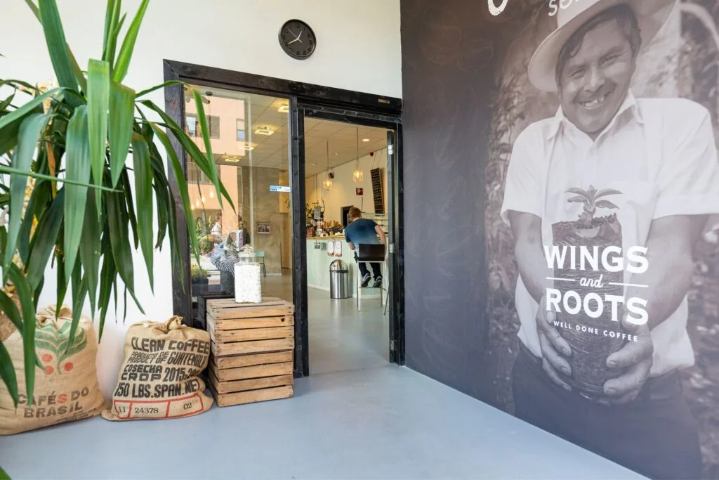 Ingang van koffiebar Langekleiweg met zicht op het interieur, koffiezakken en een grote muurfoto met de tekst "Wings and Roots".