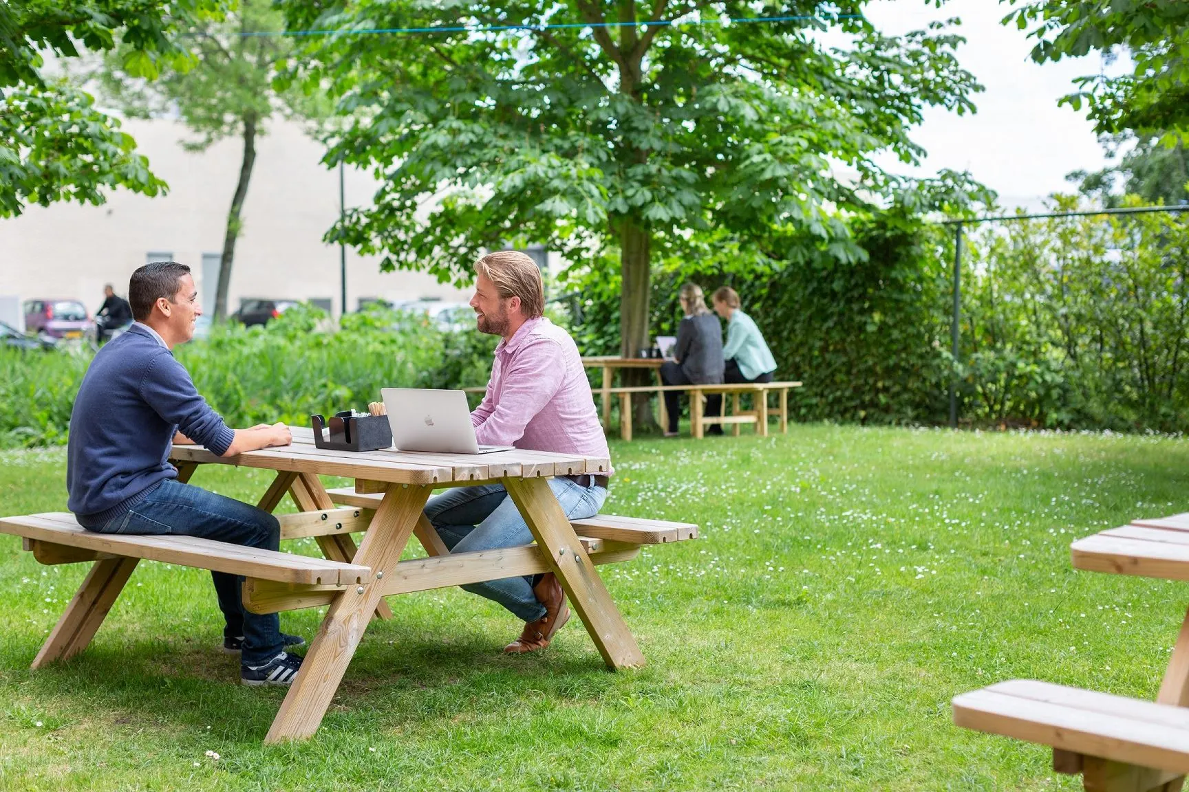 Twee mannen zitten bij Langekleiweg 9 buiten aan een picknicktafel in gesprek met een laptop op tafel.