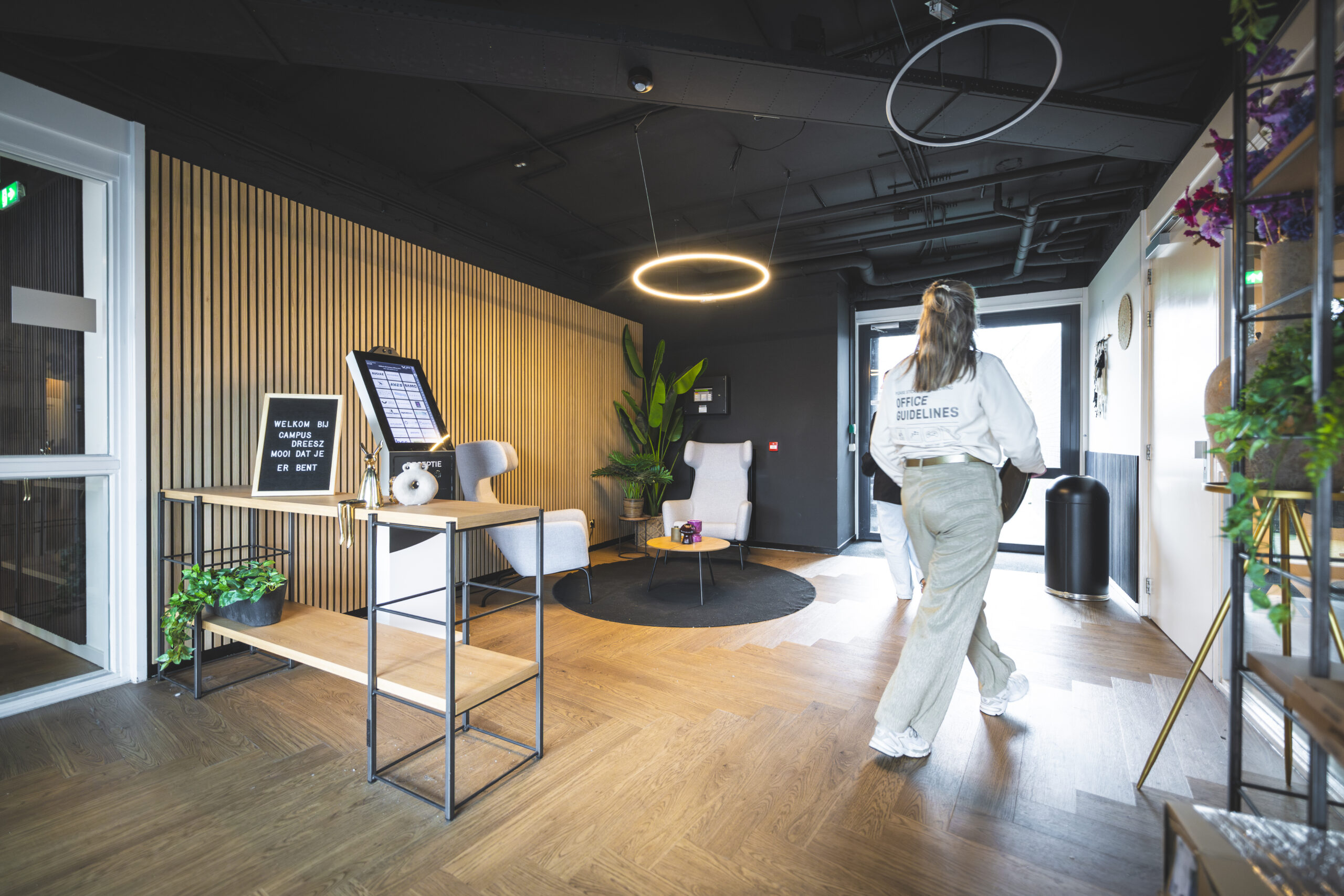 Twee mensen lopen een modern ingerichte ontvangsthal binnen met houten vloer en zitgedeelte.