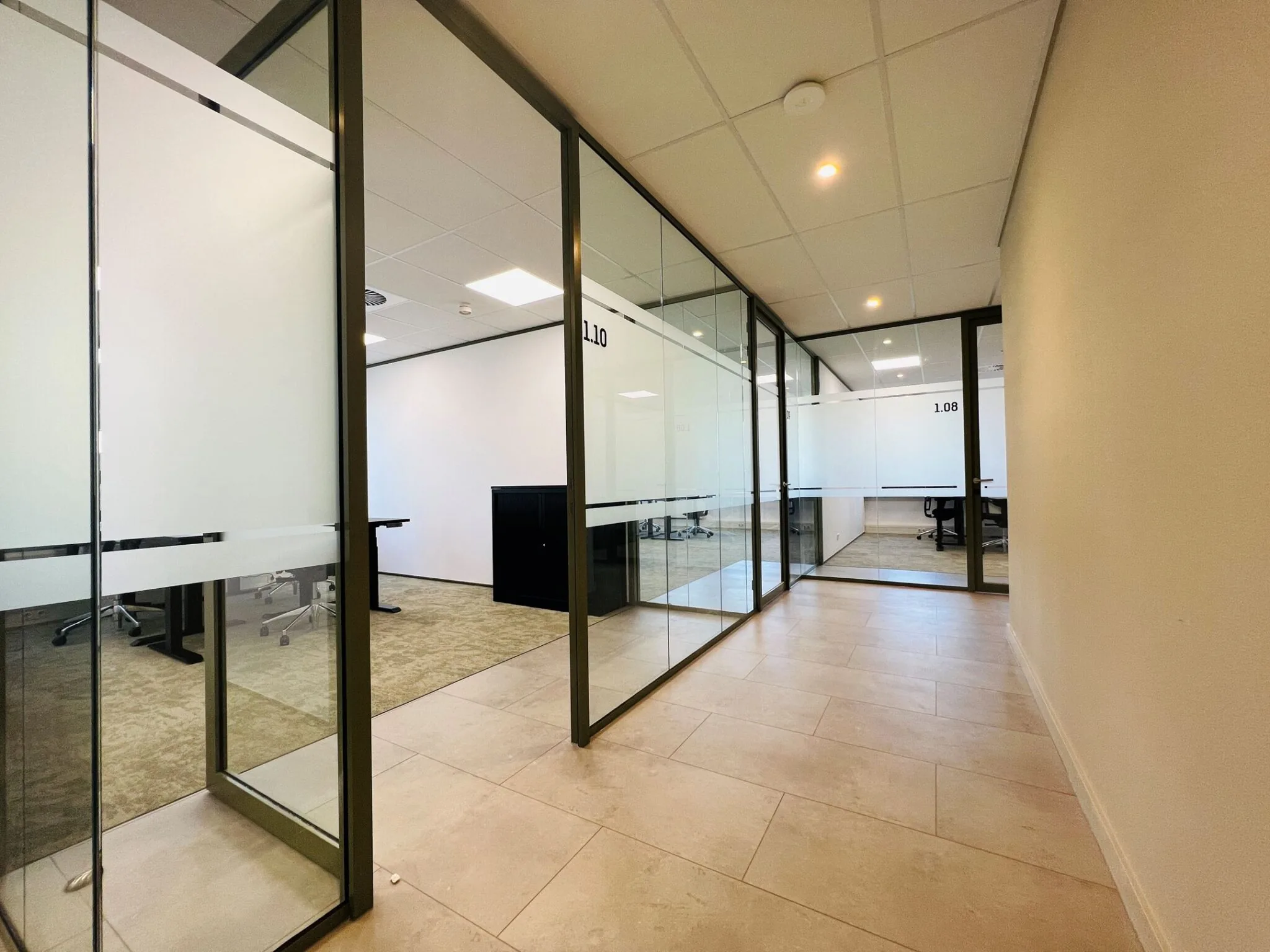 Hal met glazen kantoorkamers in een modern kantoor, waaronder ruimtes 1.08 en 1.10.