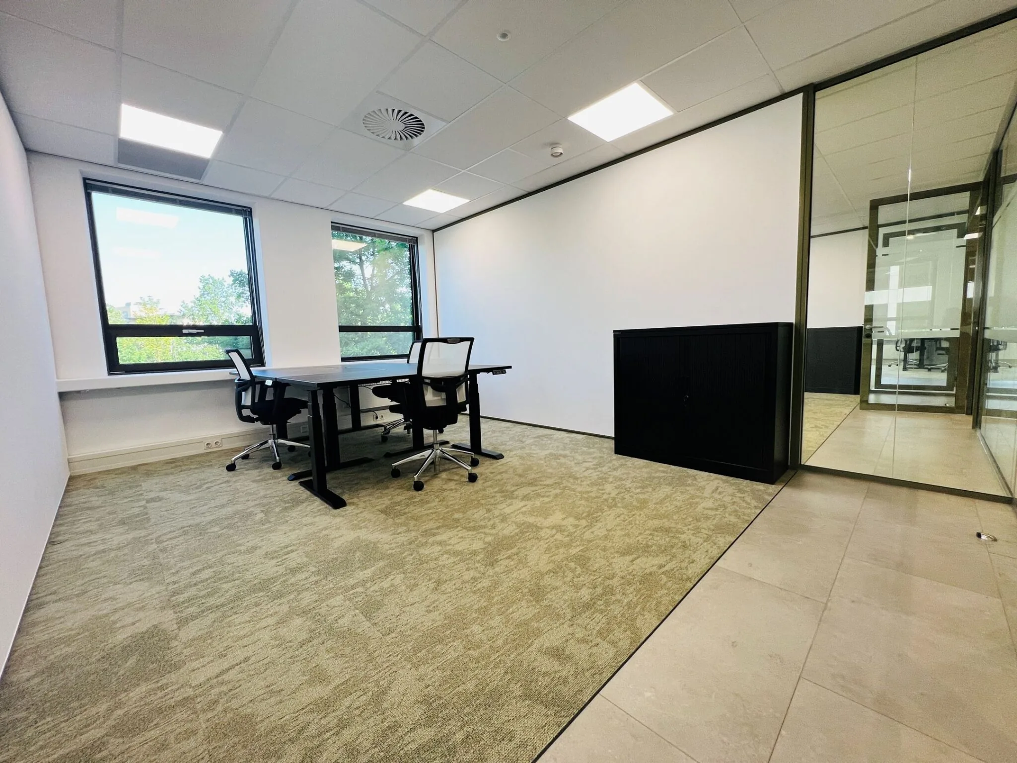 Modern kantoor met twee bureaus, bureaustoelen, grote ramen en glazen scheidingswand.