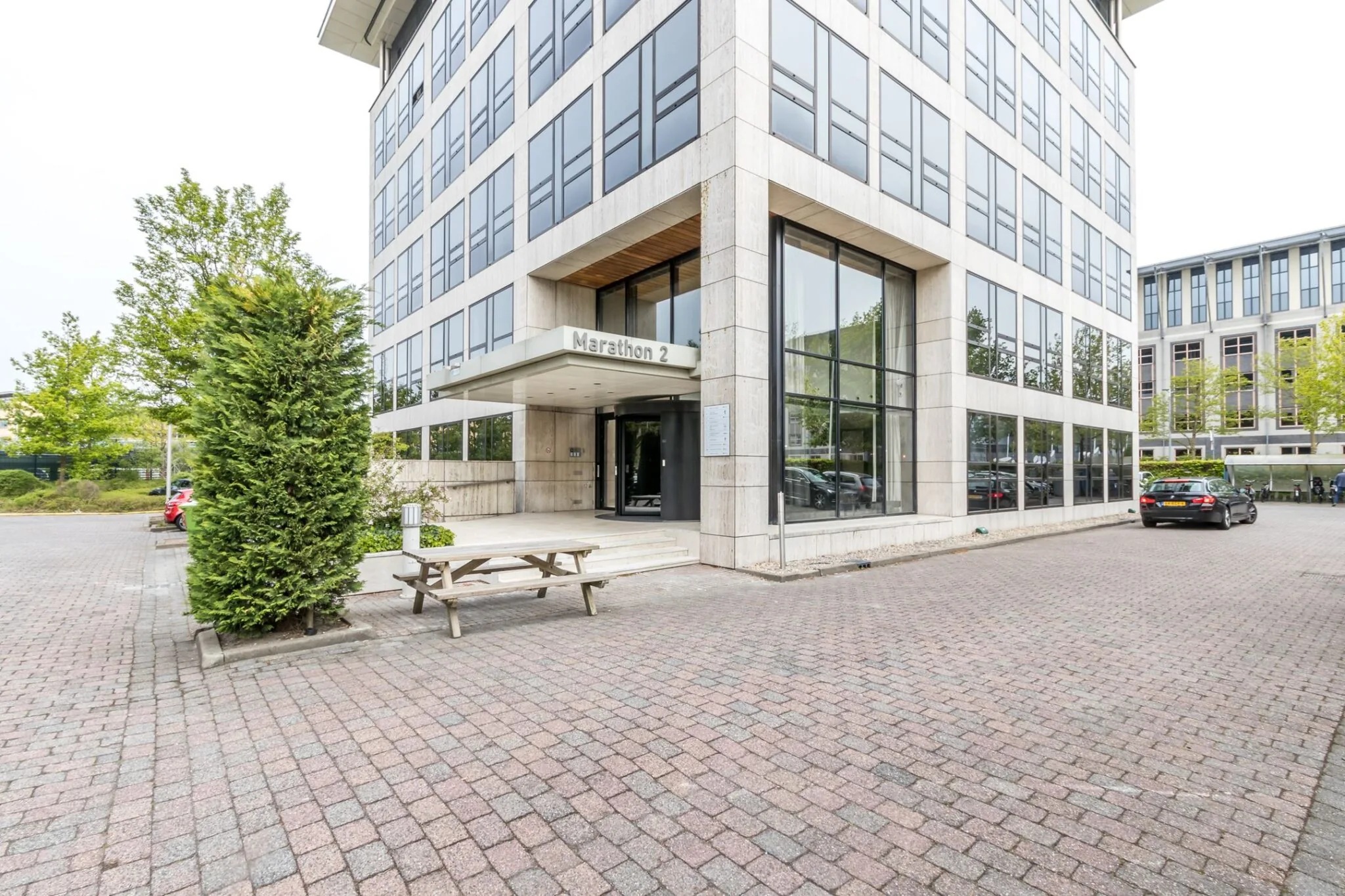 Ingang van het kantoorgebouw Marathon 2 met grote ramen, een zitbank en een geparkeerde auto op de voorgrond.