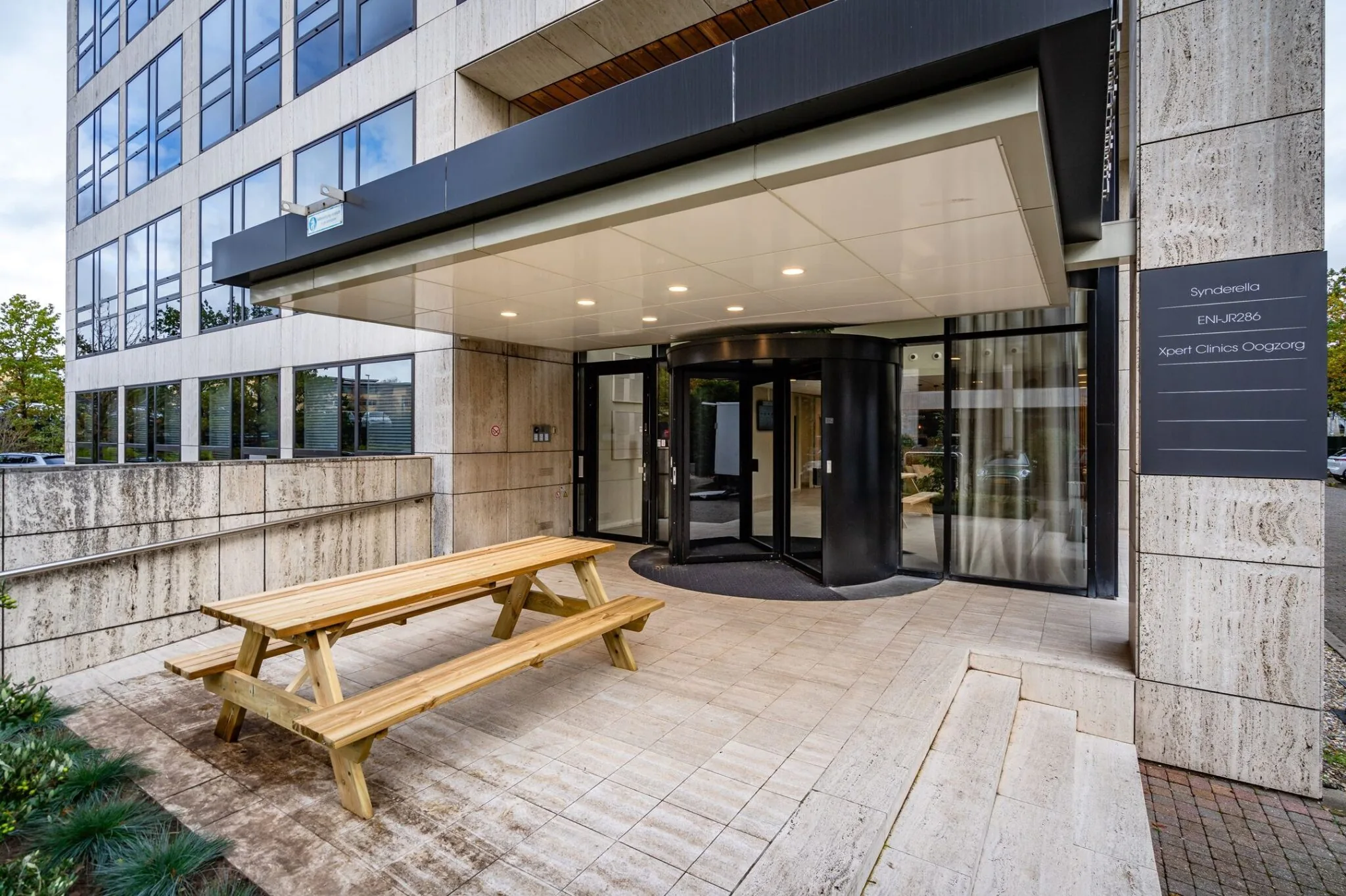 Ingang van een modern kantoorpand met een draaideur en een houten picknicktafel op het terras.