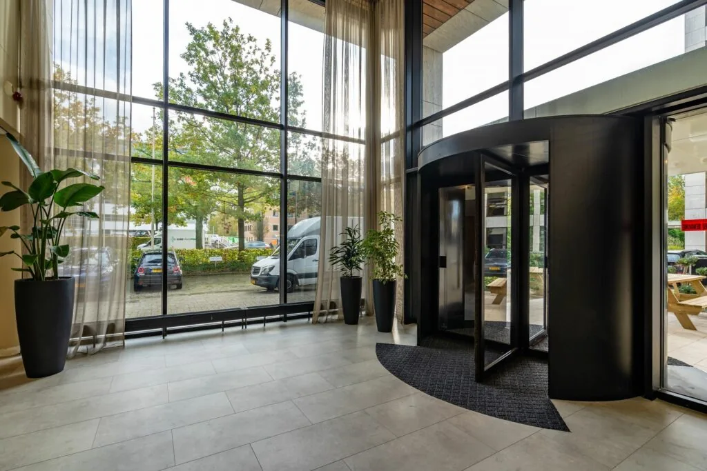 Entreehal van kantoorpand Marathon 2 met grote raampartijen, draaideur en zicht op geparkeerde auto's buiten.