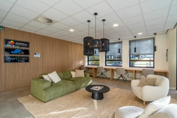 Moderne wachtruimte met groene bank, werkplekken bij het raam en logo’s van bedrijven aan een houten wand.