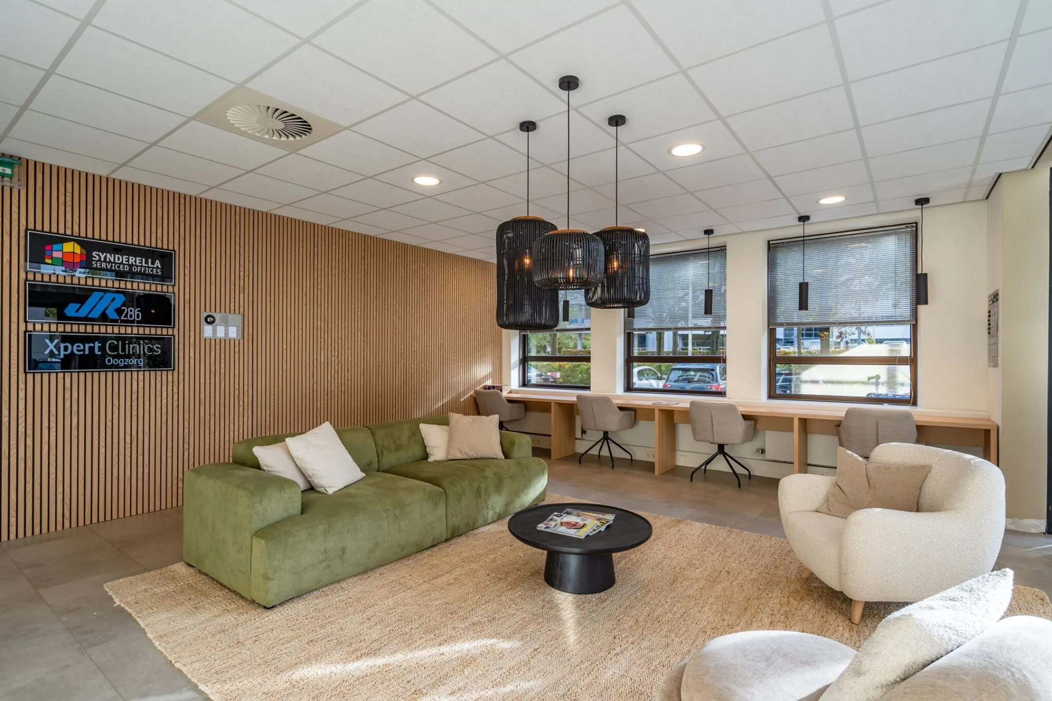 Moderne wachtruimte met groene bank, werkplekken bij het raam en logo’s van bedrijven aan een houten wand.
