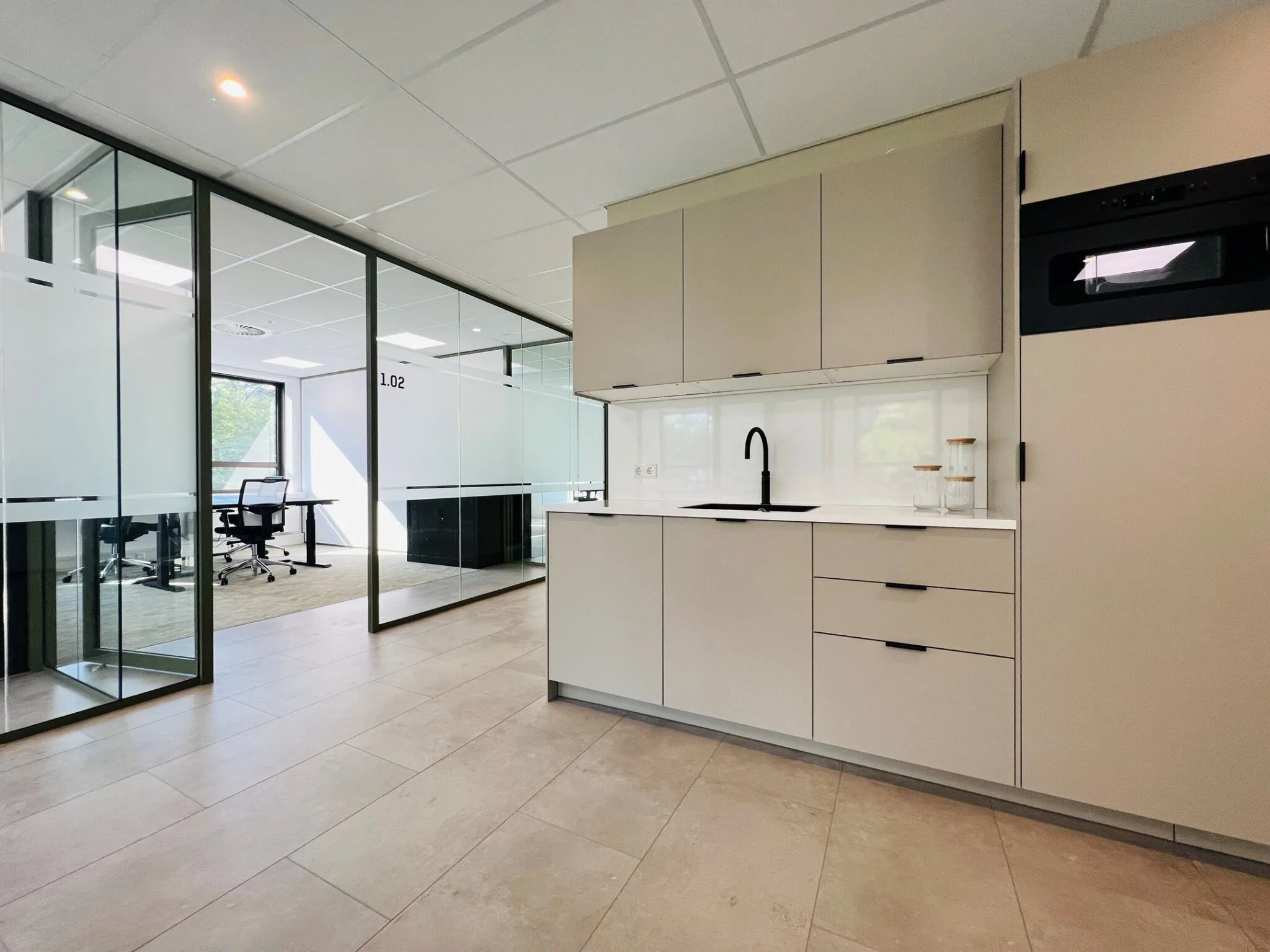 Moderne kantoorruimte met glazen wanden en een kitchenette met beige kasten en een zwarte kraan.
