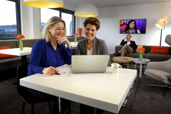 Twee vrouwen werken samen aan een laptop in een moderne lounge op Marshalllaan 2, met een ontspannen zithoek op de achtergrond.
