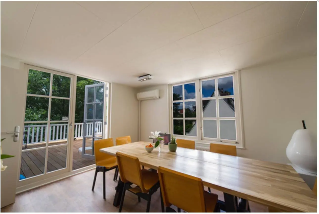 Lichte vergaderruimte met houten tafel, gele stoelen en uitzicht op een balkon en tuin via grote ramen.