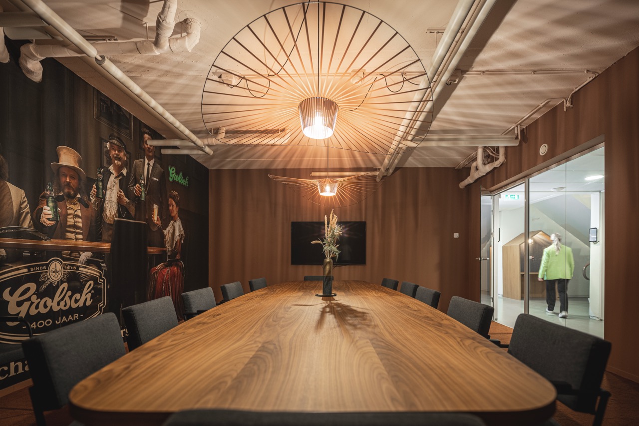 Vergaderruimte Meester met een groot houten tafel, moderne verlichting, een wanddecoratie van Grolsch en een scherm aan de muur.