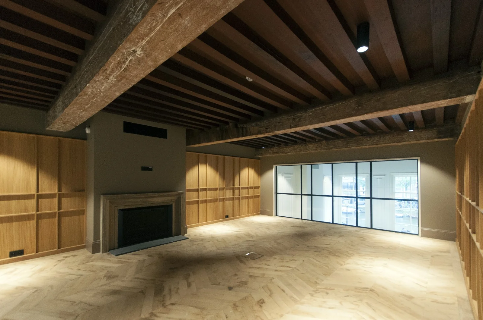 Vergaderruimte met houten vloer, houten plafondbalken, ingebouwde open haard en glazen wand met uitzicht naar buiten.