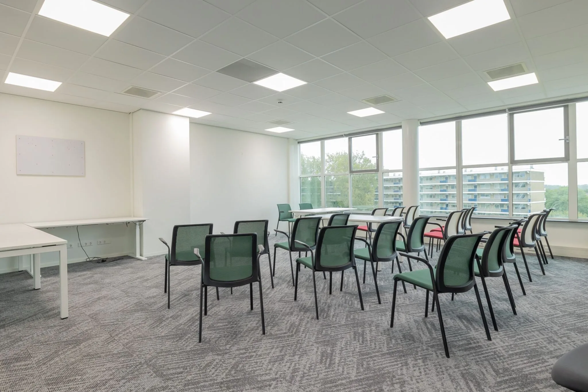 Vergaderruimte met opgestelde stoelen en tafels bij Mendelweg 32, met grote ramen en uitzicht op omliggende flatgebouwen.