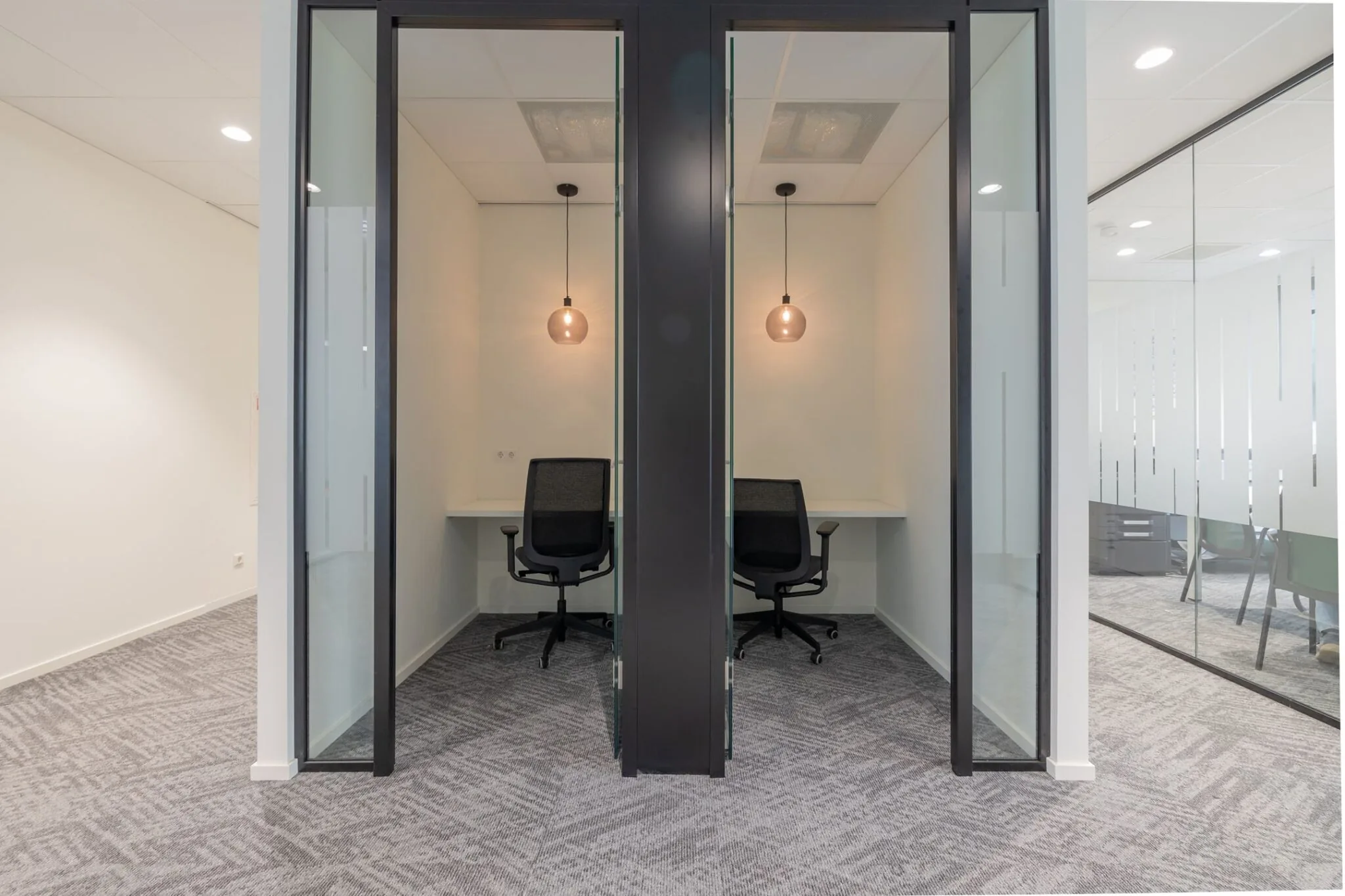 Twee moderne, glazen belcellen met bureaustoelen en hanglampen in een kantoorpand aan de Mendelweg 32.