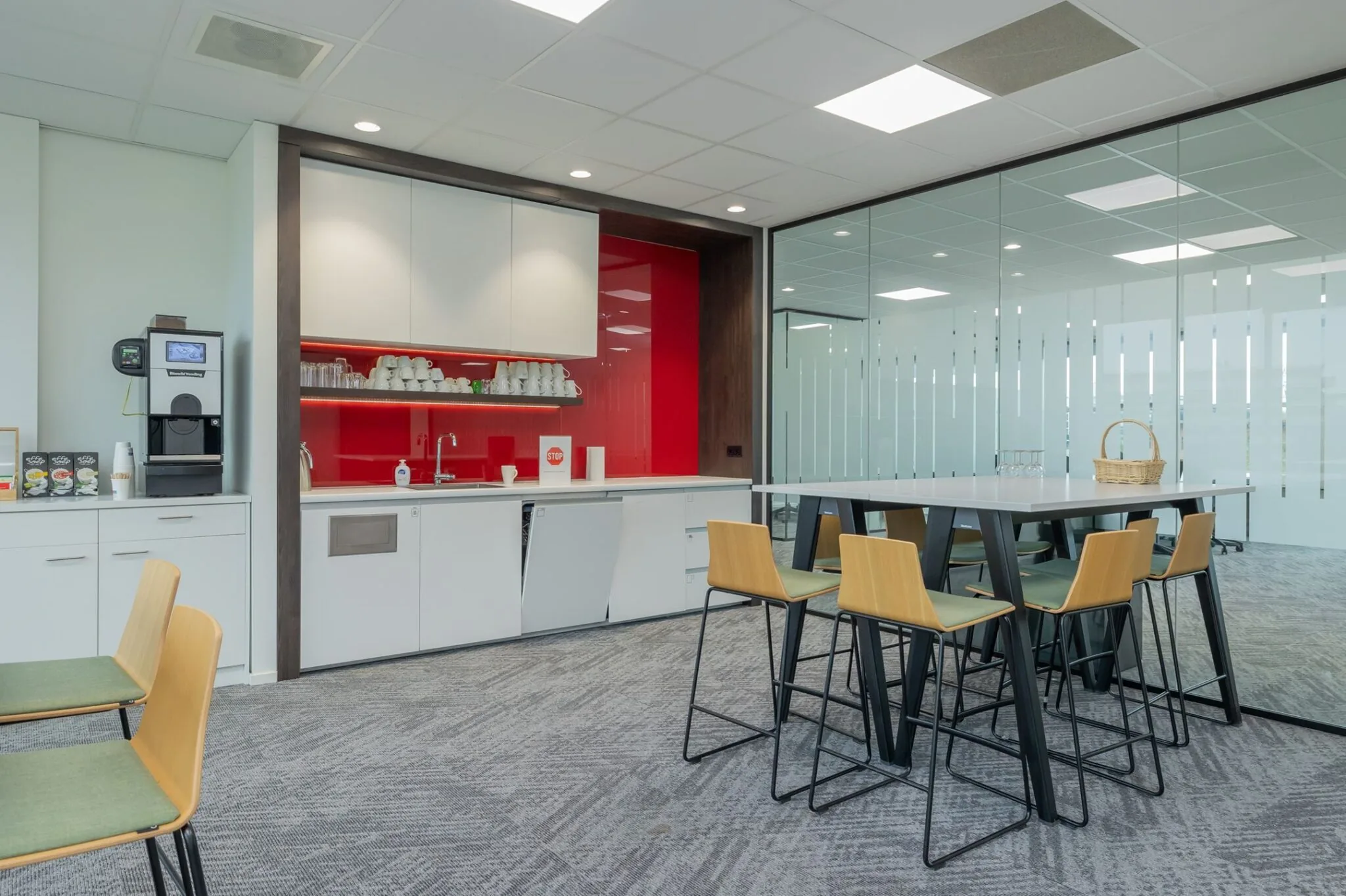 Moderne kantoorpantry met koffieautomaat, witte keukenkasten, rode achterwand en hoge vergadertafel met barkrukken.