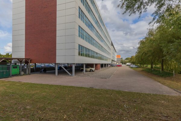 Langwerpig kantoorgebouw aan de Mendelweg 32 met een parkeergarage op de begane grond en een omliggend parkeerterrein met bomen.