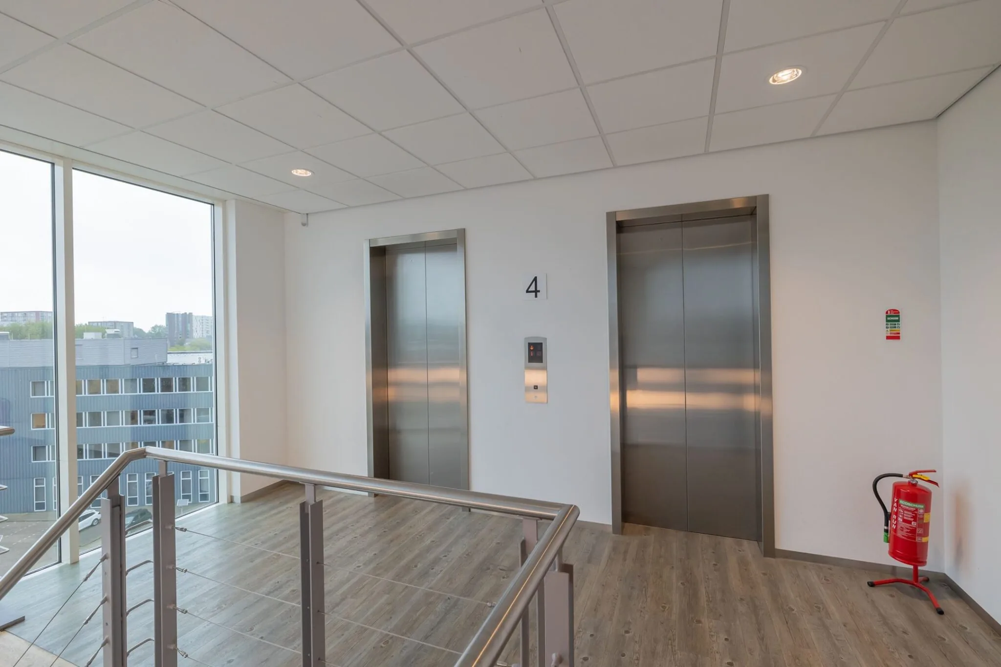 Liftlobby op de vierde verdieping van Mendelweg 32 met twee liften, grote ramen en een brandblusser.