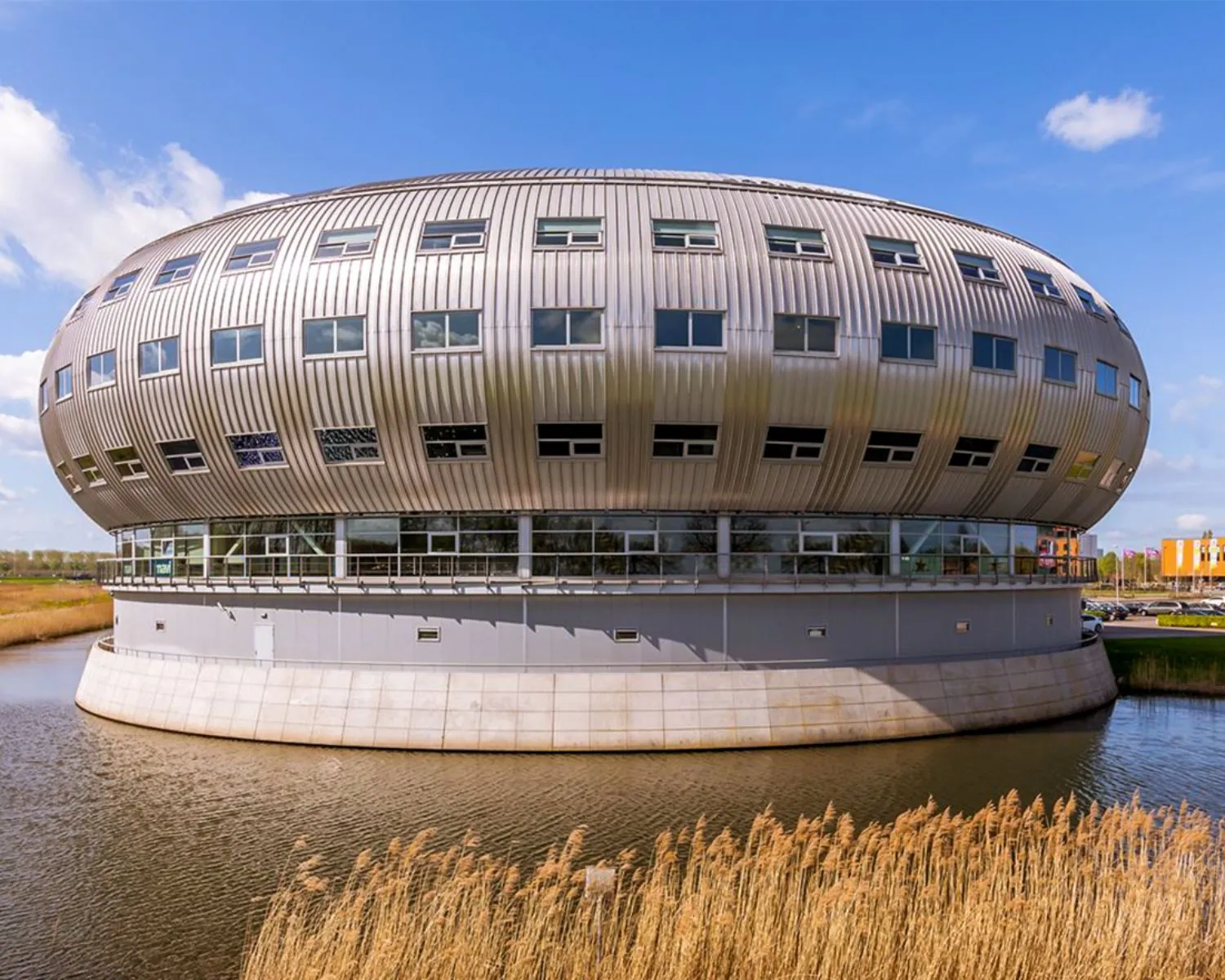 Rond, modern kantoorgebouw MyOffice Almere met metalen gevel, omringd door water.