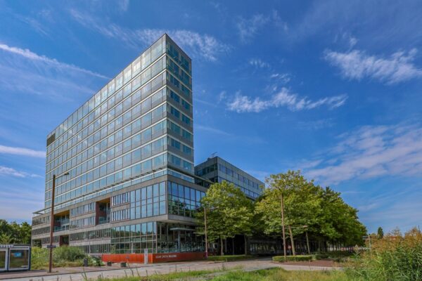 Een modern kantoorgebouw met de naam "Adam Smith Building" in Amsterdam omgeven door bomen en een blauwe lucht.