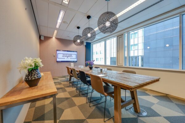 Vergaderruimte met een lange houten tafel, moderne hanglampen en een scherm met de tekst "We Go Where The Business Is".