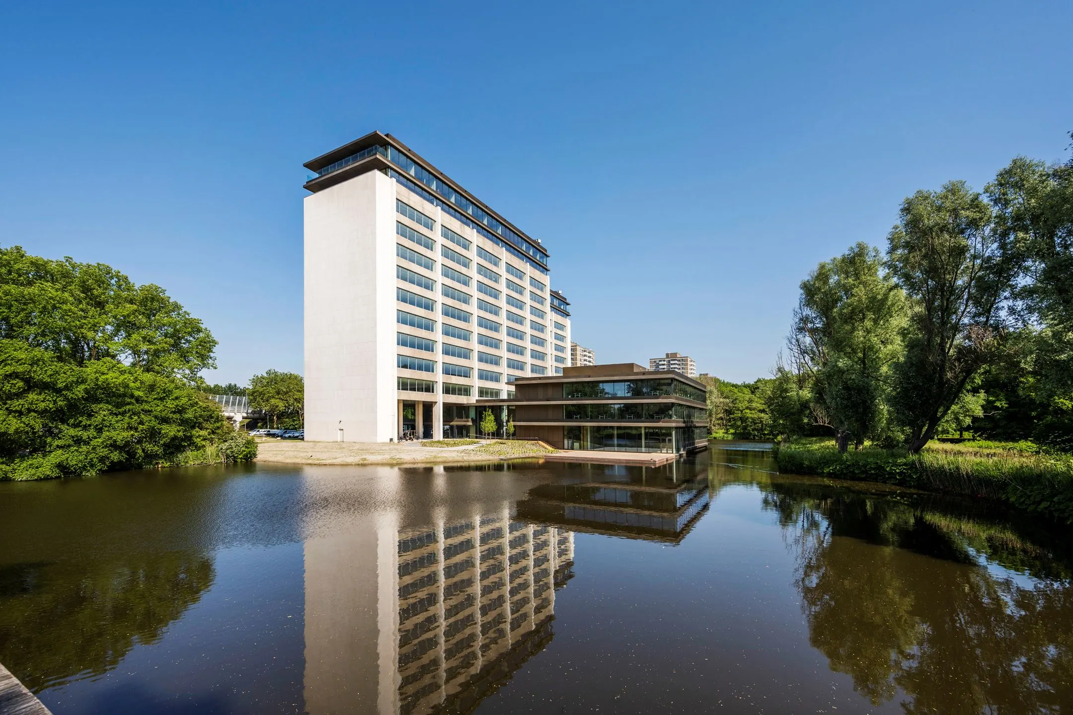 Modern kantoorgebouw aan de Nachtwachtlaan 20 met spiegeling in het naastgelegen water onder een heldere blauwe lucht.