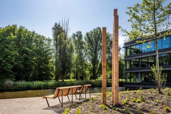Bankjes aan het water bij een modern kantoorgebouw aan de Nachtwachtlaan, omgeven door bomen en kunstzinnige houten palen.