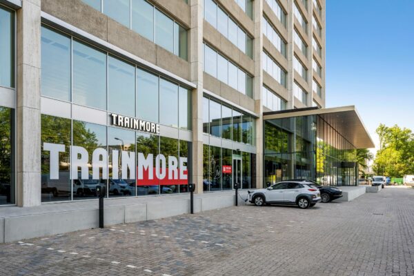 Vooraanzicht van het gebouw aan de Nachtwachtlaan 20 met de ingang van sportschool TrainMore en geparkeerde auto’s op de oprit.