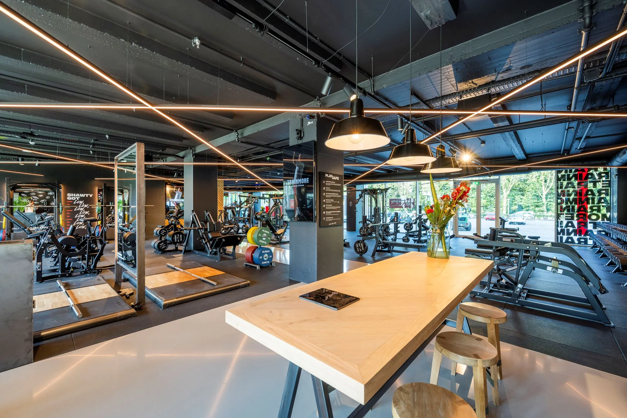 Moderne fitnessruimte aan de Nachtwachtlaan 20 met krachttrainingsapparatuur, halterplatforms en een zithoek met houten tafel en bloemenvaas.