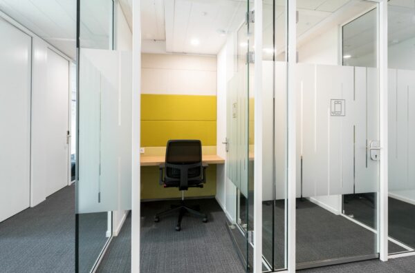 Kleine glazen belruimte met een bureau, bureaustoel en gele wandbekleding in Nevelgaarde 8.