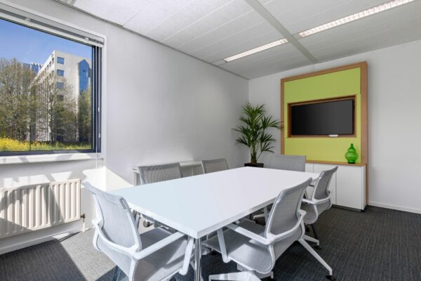 Vergaderruimte in Nevelgaarde 8 met witte tafel, zes stoelen, flatscreen, plant en uitzicht op kantoorpand.