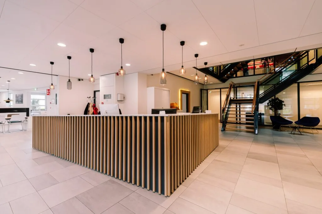 Moderne ontvangsthal met houten receptiebalie, hanglampen en een trap naar de bovenverdieping in het kantoorgebouw aan de Newtonlaan 115.