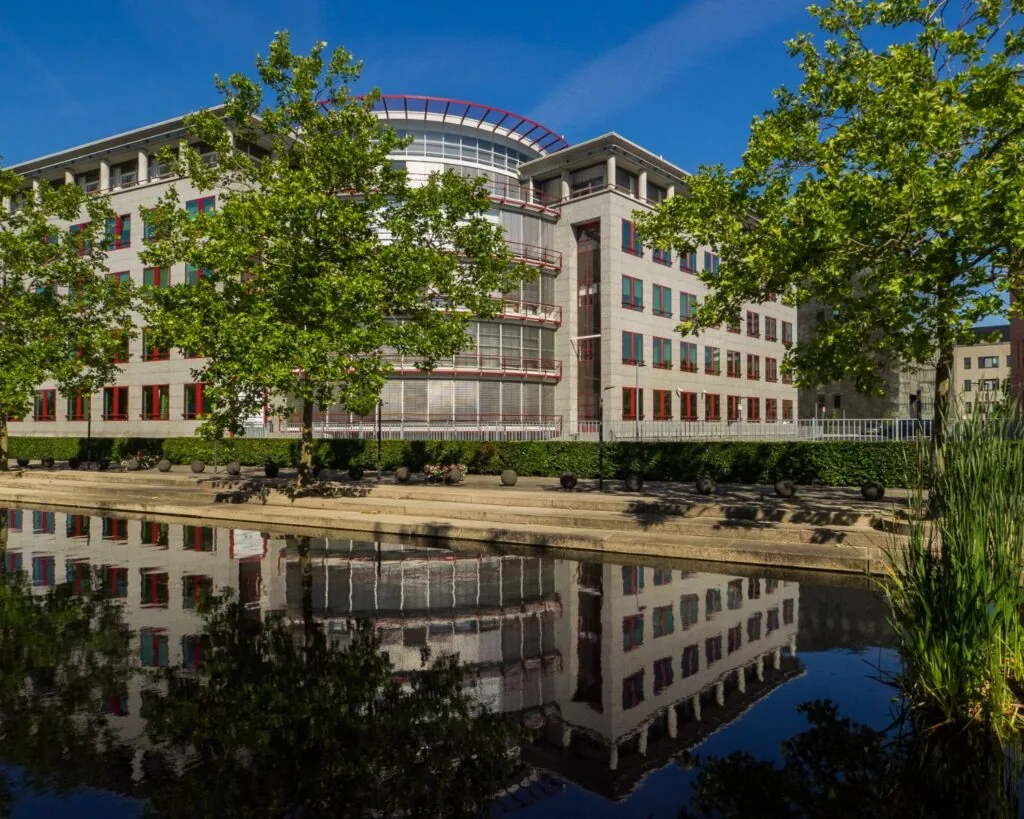 Kantoorgebouw aan de Newtonlaan 51-97 in Utrecht, weerspiegeld in een vijver met groene bomen op de voorgrond.