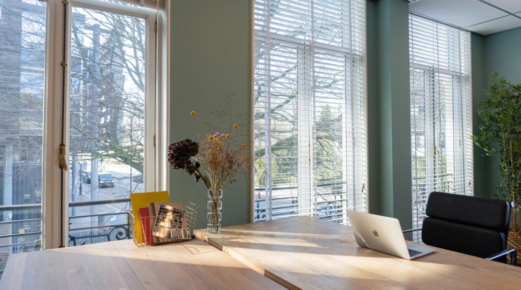 Licht en modern kantoorinterieur in Office Hotel Kennemerplein met houten bureau, laptop en grote ramen met jaloezieën.